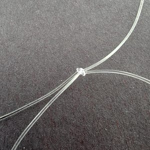 K&ouml;nnte beinhalten: Nahaufnahme eines klaren, d&uuml;nnen, transparenten Materials, das zu einem Knoten gebunden ist. Das Material ist gekr&uuml;mmt und &uuml;ber eine dunkelgraue Oberfl&auml;che abgewinkelt. Der Knoten befindet sich in der Mitte des Bildes.