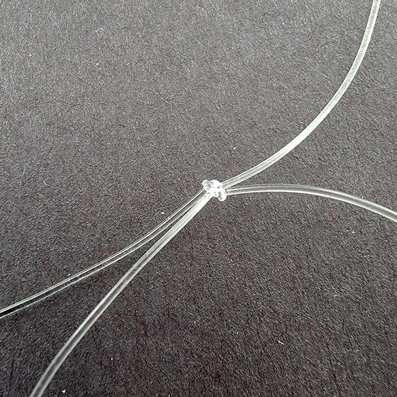 K&ouml;nnte beinhalten: Nahaufnahme eines klaren, d&uuml;nnen, transparenten Materials, das zu einem Knoten gebunden ist. Das Material ist gekr&uuml;mmt und &uuml;ber eine dunkelgraue Oberfl&auml;che abgewinkelt. Der Knoten befindet sich in der Mitte des Bildes.