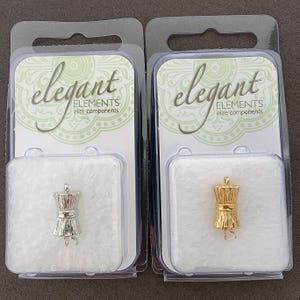 May include: Two clear plastic packaging displays with white backgrounds and gold and silver metal charms. The charms are shaped like a bundle of wheat. The packaging has the text "elegant elements elite components".