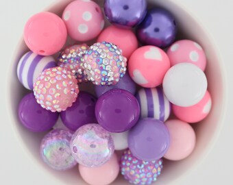 May include: A collection of colorful beads in a white bowl. The beads are various shades of pink, purple, and white, with some featuring polka dots, stripes, and sparkling embellishments. These beads are perfect for jewelry making and crafts.