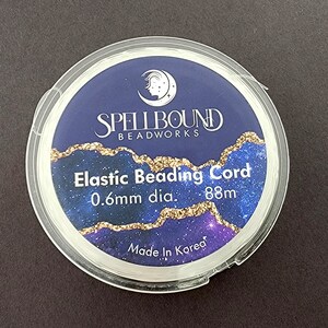 K&ouml;nnte beinhalten: Eine transparente Kunststoffspule mit elastischer Schnur zum Auff&auml;deln von Perlen. Das Etikett tr&auml;gt die Aufschrift "Spellbound Beadworks" mit dem Text "Elastic Beading Cord 0.6mm dia. 88m Made in Korea". Der Hintergrund ist dunkelblau mit goldenen Akzenten.