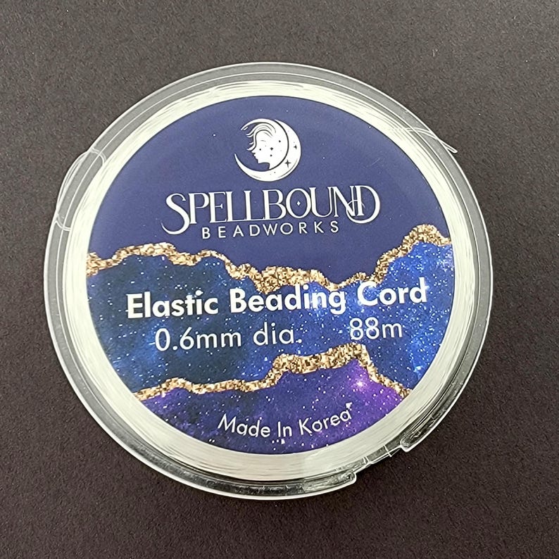 K&ouml;nnte beinhalten: Eine transparente Kunststoffspule mit elastischer Schnur zum Auff&auml;deln von Perlen. Das Etikett tr&auml;gt die Aufschrift "Spellbound Beadworks" mit dem Text "Elastic Beading Cord 0.6mm dia. 88m Made in Korea". Der Hintergrund ist dunkelblau mit goldenen Akzenten.