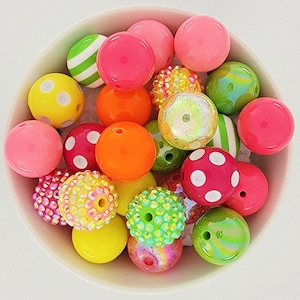 May include: A collection of colorful round beads in a white bowl. The beads are various colors, including pink, orange, yellow, green, and red. Some beads have patterns like polka dots and stripes. Others are covered in small, sparkling embellishments. These beads are perfect for jewelry making.