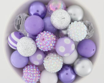 May include: A bowl filled with a variety of purple, white, and silver beads. The beads are round and have different textures, including smooth, sparkly, and textured.