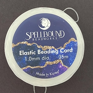 K&ouml;nnte beinhalten: Eine Spule mit transparentem, elastischem Schmuckband. Das Etikett ist dunkelblau mit goldenen Glitzerakzenten und dem Text "Spellbound Beadworks". Der Text "Elastic Beading Cord" und "1,0 mm dia." sind ebenfalls sichtbar. Die Spule enth&auml;lt 35 m Band und ist in Korea hergestellt.