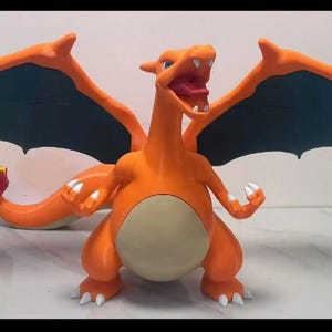 Könnte beinhalten: Eine orangefarbene Charizard-Figur mit ausgebreiteten Flügeln und einer Flamme am Schwanz. Die Figur hat einen hellbeige Bauch, weiße Krallen und eine rote Zunge. Die Flügel sind orange mit einer dunkelgrünen Unterseite. Sammlerstück.