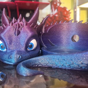 May include: A 3D printed dragon figurine with a purple and blue iridescent finish. The dragon has a curled tail and a small, open mouth.