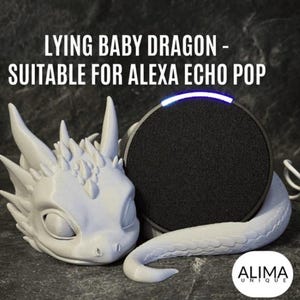 Op de afbeelding: Een wit 3D-geprint baby-drakenbeeldje met een lichtblauwe gloed rond de luidspreker. De draak ligt met zijn kop op een zwarte Alexa Echo Pop slimme luidspreker. De tekst "LYING BABY DRAGON - SUITABLE FOR ALEXA ECHO POP" staat boven de afbeelding. De tekst "ALIMA UNIQUE" staat onder de afbeelding.