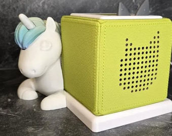 Puede incluir: Una figura de unicornio blanco con melena y cuerno azul y verde descansa junto a un altavoz verde lima. El altavoz tiene un diseño perforado y se asienta sobre una base blanca. El altavoz tiene dos orejas grises.