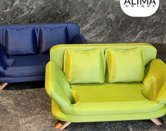 Puede incluir: Dos sofás en miniatura, uno azul y otro verde, con cojines. Los sofás están hechos de un material suave y mullido y tienen una forma redondeada. Los sofás están sobre una superficie gris. El texto "ALIMA UNIQUE" es visible en la esquina superior derecha de la imagen.