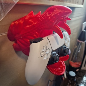 Könnte beinhalten: Ein roter, 3D-gedruckter Drachenkopf-Wandhalter mit einem weißen und schwarzen Videospiel-Controller im Maul.