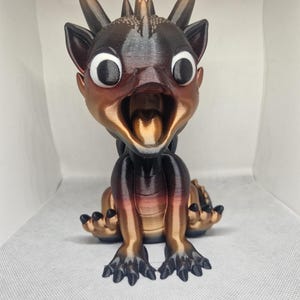 Könnte beinhalten: Eine kleine, 3D-gedruckte Drachenfigur mit einer schwarzen und kupferfarbenen Metallic-Oberfläche. Der Drache hat große, runde Augen und einen offenen Mund, der eine kupferfarbene Zunge zeigt. Der Drache sitzt auf seinen Hinterbeinen mit ausgestreckten Vorderpfoten.