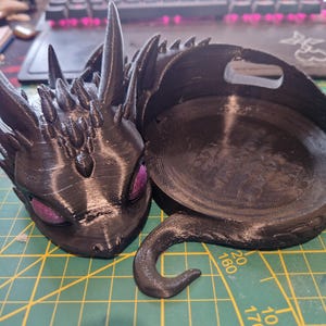 Peut inclure: Une figurine de dragon noir imprimée en 3D avec des yeux violets. Le dragon est enroulé dans une position de sommeil avec sa queue enroulée autour de son corps. Le dragon est conçu pour être un support de téléphone.