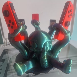 Può includere: Un supporto a forma di polpo stampato in 3D con una finitura iridescente turchese tiene due Joy-Con rossi Nintendo Switch.