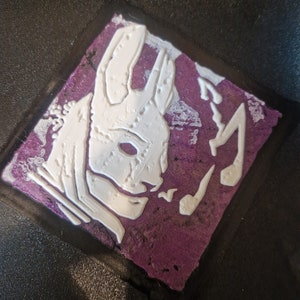 Könnte beinhalten: Ein weißes, 3D-gedrucktes Emblem eines Kaninchenkopfes auf einem violetten Hintergrund. Das Kaninchen hat ein schädelartiges Aussehen mit einer großen, hervorstehenden Nase und scharfen Zähnen.