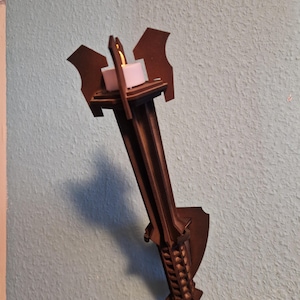 Op de afbeelding: Een bruine houten wandkandelaar met een kaarsenhouder. De kandelaar heeft een decoratief ontwerp met een schildvormige achterplaat en een fakkelachtig uiterlijk. De kaars is wit.