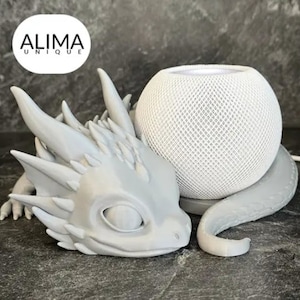 May include: A grey 3D printed dragon figurine holding a white smart speaker. The dragon has a detailed face with a closed eye and a textured body. The speaker has a mesh covering and a white base.