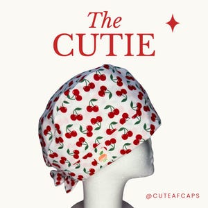 May include: A white scrub cap featuring a repeating pattern of red cherries with green stems. The text The CUTIE is printed in red above the cap, and the text @CUTEAFCAPS is printed in red below. A small white label with an orange icon is visible.