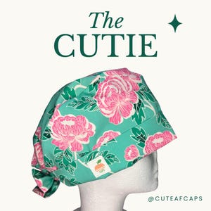 May include: A teal surgical scrub cap with a floral pattern of pink peonies and green leaves. The cap is labeled "The Cutie" in dark green text. A small white tag with an orange logo is visible.