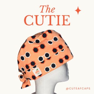 May include: A peach-colored surgical scrub cap with a repeating pattern of sunglasses. The text "The CUTIE" is displayed in orange, with a small logo patch. The cap is tied at the back.
