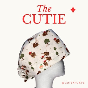 May include: A white surgical scrub cap featuring a repeating pattern of brown Highland cows with red bows, trees, and oil derricks. The text "The CUTIE" is in red, with the brand name "Cute AF Caps" visible.