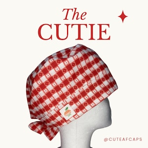 May include: A red and white gingham scrub cap featuring a small white flower pattern within the squares. The cap includes a small white rectangular tag with an orange peach graphic and the text CUTE AF CAPS. The text The CUTIE is above.