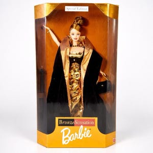 Puede incluir: Una muñeca Barbie en una caja de plástico transparente. La muñeca lleva un vestido dorado y negro y una capa negra. La caja tiene las palabras "Bronze Sensation" y "Special Edition" impresas.