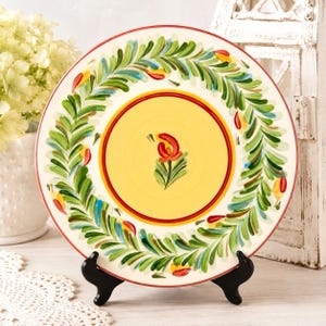 May include: A round ceramic plate featuring a central yellow circle with a single red flower motif, surrounded by a wide border of hand-painted green leafy wreaths and small red and yellow accents, resting on a black metal display stand.