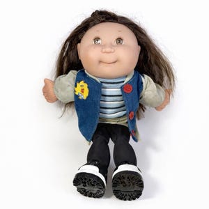 May include: A vintage doll with brown hair, freckles, and a smiling expression. The doll wears a denim vest with a yellow patch, a striped shirt, black pants, and black and white shoes. The doll is a collectible item.