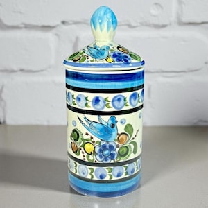 Puede incluir: Un tarro de cerámica con tapa, con un diseño pintado a mano. El tarro es cilíndrico con una combinación de colores azul y blanco, adornado con imágenes de pájaros azules, flores y bayas. La tapa tiene un diseño a juego y un adorno azul.