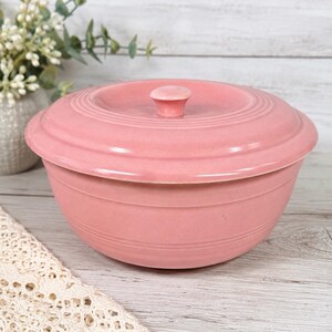May include: A round, dusty pink ceramic bowl with a matching lid. The bowl features a smooth, glazed finish and subtle horizontal ring detailing around the exterior. A small, centered knob handle sits atop the lid, completing the design.
