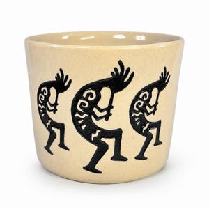 May include: A beige ceramic planter featuring three black silhouettes of Kokopelli figures playing flutes. The cylindrical vessel has a smooth, speckled finish and a wide opening, showcasing the dark, detailed graphic design against the light base.