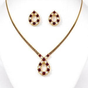 Könnte beinhalten: Ein goldfarbenes Schmuckset mit einer Halskette und passenden Ohrringen. Die Halskette hat einen tropfenförmigen Anhänger und die Ohrringe sind tropfenförmig. Sowohl der Anhänger als auch die Ohrringe sind mit roten und klaren Edelsteinen besetzt.