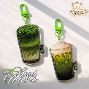 May include: Two acrylic keychains featuring iced matcha drinks. One shows a layered matcha drink with ice cubes, the other with a foamy topping. Both have green clasps and the text "抹茶" in a small black rectangle. The words "Iced Matcha" are written in cursive.