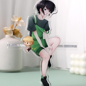 May include: A clear acrylic standee featuring an anime-style character with black hair wearing a dark green t-shirt and green overalls with a small frog graphic. A smaller, chibi-style character with blonde hair sits beside the main figure.