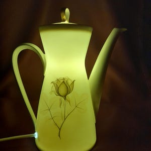 Könnte beinhalten: Eine leuchtende, cremefarbene Keramik-Teekannenlampe mit floralem Design. Die Lampe hat einen gebogenen Griff, einen langen Ausguss und einen Deckel mit einem goldfarbenen Griff. Das florale Design ist eine Rose mit Blättern. Die Lampe ist eingesteckt und beleuchtet.