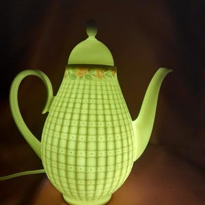 Może przedstawiać: Świecący zielony ceramiczny imbryk z wzorem kratki i kwiatowym wzorem na obrzeżu. Imbryk ma uchwyt, dzióbek i pokrywkę z małym guzikiem. Wewnętrzne oświetlenie tworzy miękki, ciepły blask.