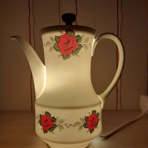 Puede incluir: Lámpara eléctrica con forma de tetera de color crema, con tapa y asa negras. La lámpara presenta diseños florales de rosas rojas con hojas verdes. La lámpara se ilumina desde el interior, creando un brillo suave.