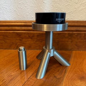 May include: A silver metal stand with three legs and a round top. A black Amazon Echo Dot is on top of the stand. A silver metal extension piece is on the floor next to the stand.