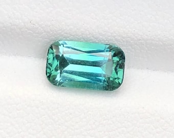 May include: A rectangular, emerald-cut gemstone with a gradient of teal and green hues. The gem is set against a white background, showcasing its clarity and color. The facets create a play of light, highlighting the gem's brilliance.