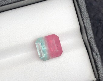 May include: A rectangular gemstone with a bi-color design, featuring a blend of pink and teal hues. The gem is set within a black box with a white interior, creating a striking contrast. The stone's facets reflect light, showcasing its clarity and cut.