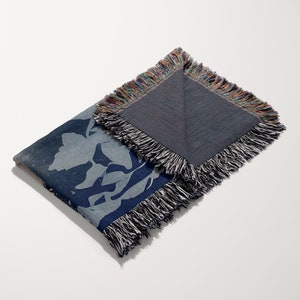 May include: A blue woven throw blanket with a floral pattern and fringed edges. The blanket is folded in half and the back side is visible.