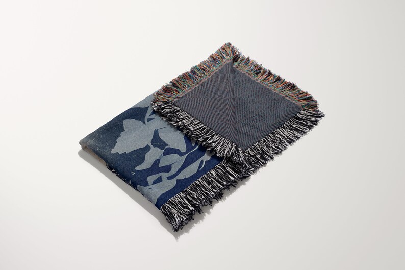 May include: A blue woven throw blanket with a floral pattern and fringed edges. The blanket is folded in half and the back side is visible.