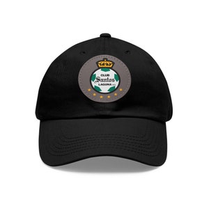 Puede incluir: Gorra de béisbol negra con un parche bordado en verde, blanco y dorado con el texto "Club Santos Laguna" y un balón de fútbol.