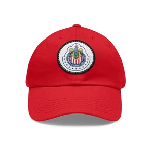 Puede incluir: Gorra de béisbol roja con un parche bordado blanco y azul con el logotipo del Club Deportivo Guadalajara, un equipo de fútbol profesional mexicano.