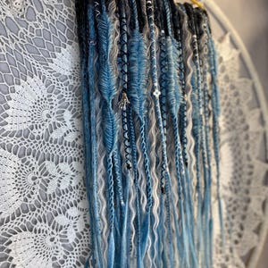 May include: A collection of blue, gray, and black synthetic dreadlocks hanging from a gold-colored hanger. The dreadlocks are decorated with beads and charms. The background features a white, lacy, circular doily.