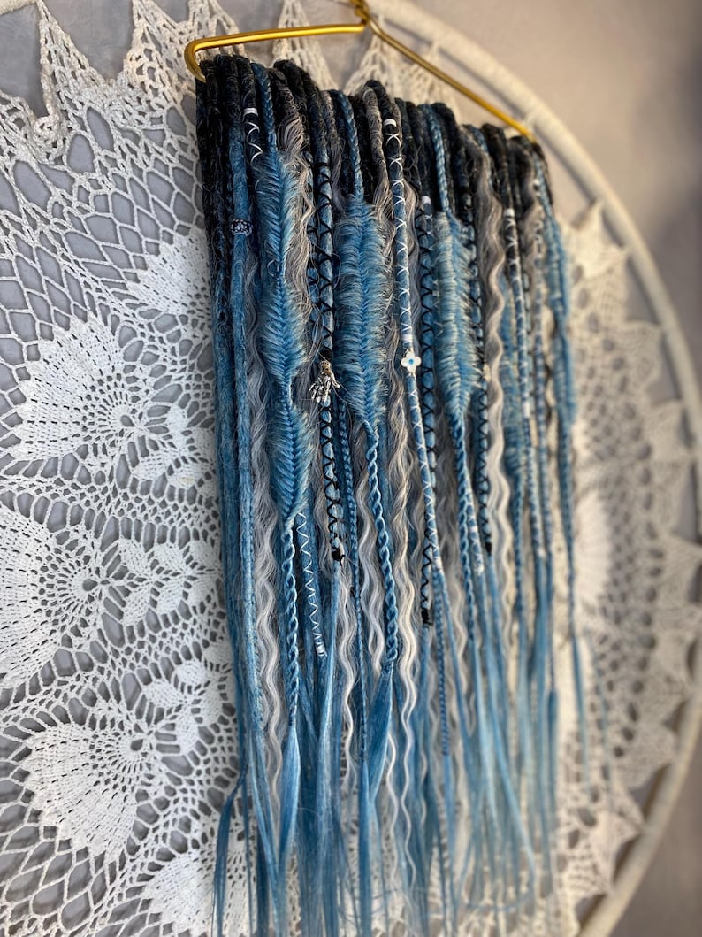 May include: A collection of blue, gray, and black synthetic dreadlocks hanging from a gold-colored hanger. The dreadlocks are decorated with beads and charms. The background features a white, lacy, circular doily.