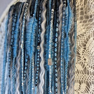 May include: A collection of blue, gray, and black dreadlocks, some with wavy textures and others braided. The dreadlocks are adorned with white and black stitching, beads, and blue feather-like accents. They hang from a gold-colored bar.