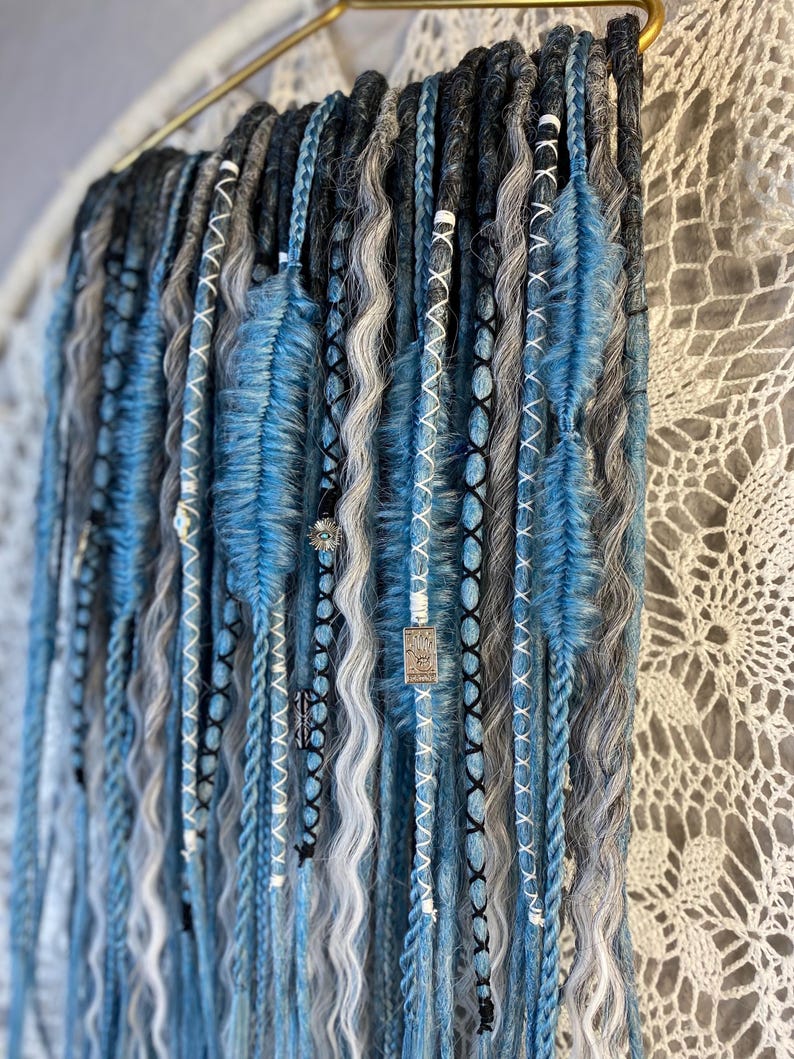 May include: A collection of blue, gray, and black dreadlocks, some with wavy textures and others braided. The dreadlocks are adorned with white and black stitching, beads, and blue feather-like accents. They hang from a gold-colored bar.