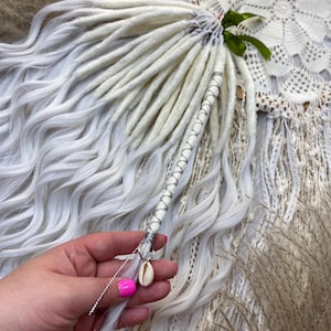 May include: A bundle of white and gray dreadlocks with a decorative wrap. The wrap is silver and white, with a small seashell charm hanging from it. The dreadlocks are wavy and have a natural look.